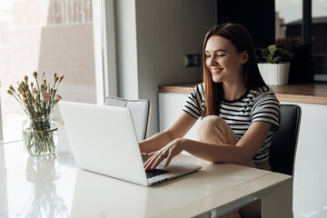 Laughing romantic glad, cheerful young brown long haired woman sitting in white kitchen studio, using laptop in morning
