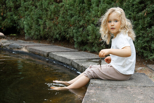 Small little blonde curly barefoot girl sitting and washing legs in lake and look at camera in park outdoors. Child care
