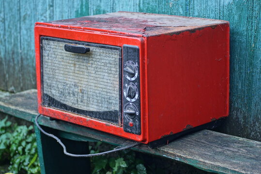 one old dirty red black metal electric oven with a gray glass door stands on a wooden table against a green wall in the street