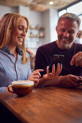 Couple Sitting At Table In Coffee Shop Looking At Mobile Phone Together