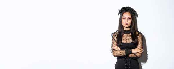 Image of beautiful asian woman in black lace dress and wreath looking serious. Girl dressed-up for halloween party as evil witch, standing over white background