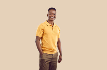 Fototapeta premium Young smiling African American man millennial with short hair posing with one hand in pocket pants and confidently looking at camera dressed in casual clothes stands on beige background. Copy space