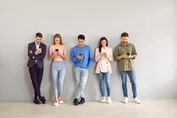 People standing in row having fun chatting on social networks without paying attention to each other. Technology-obsessed young people are smiling looking at screens of their modern mobile phones.