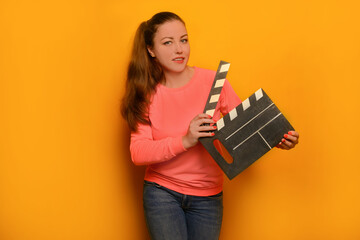 Young woman holding classic black film making clapperboard or movie slate without text and looking at camera on a yellow background.

Attractive girl wearing pink pullover or jersey and jeans.