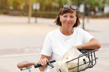 Fototapeta na wymiar Portrait of active middle ages woman on bicycle on sunny day. Mature lady in white t shirt is riding city bike in park. Leisure and lifestyle concept, smiling female cycling with her bike in summer