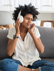 Fototapeta na wymiar Happy woman listening to music on headphones at home. Cheerful Black woman singing song with closed eyes, sitting on sofa. Hobby, relaxation, leisure activity concept