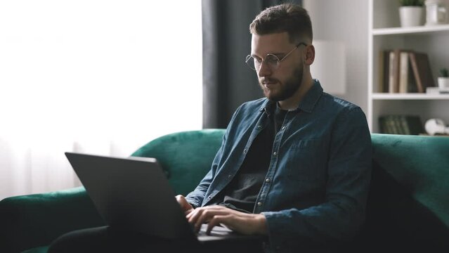 Bearded young man in denim shirt working on laptop, handsome coder writing code sitting on the sofa
