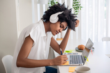 Concentrated woman in headphones writing on sticky paper. Focused African American woman sitting at computer desk and studying online during self-isolation. Online education, pandemic concept
