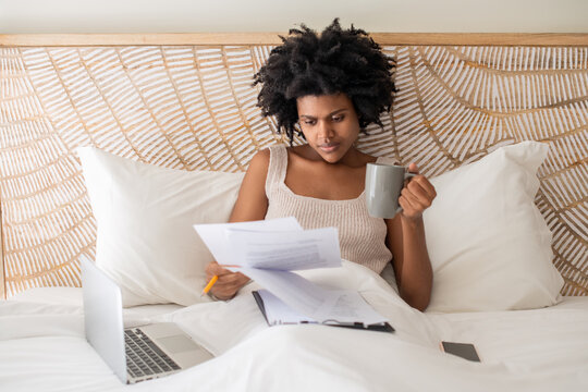 Focused freelancer lying in bed and working with papers. Serious African American woman drinking coffee working from home in morning. Home office, occupation concept