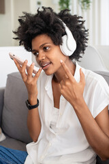 Fototapeta na wymiar Cheerful woman in headphones recording voice message. African American woman chatting with friend online, sitting on sofa. Communication, relaxation, leisure activity concept