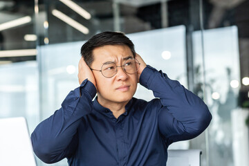 Noise in the office, Asian worker businessman covers his ears with hands from noise, man works...