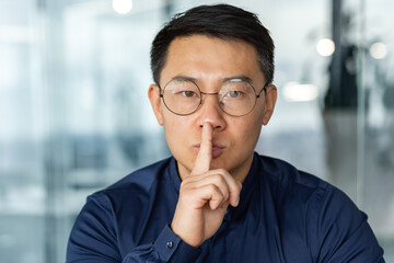Fototapeta na wymiar man in glasses and casual shirt businessman looking at camera working inside modern office building asian investor holding finger near mouth demanding silence.