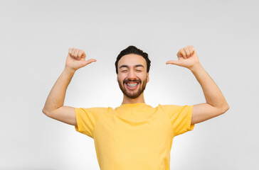 Young latin and hispanic man with beard and braces raising arms and pointing at himself with eyes closed and smiling. White background.