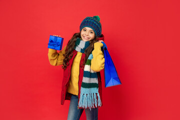 glad teen girl wearing knitted hat hold shopping bag and present box on red background, boxing day