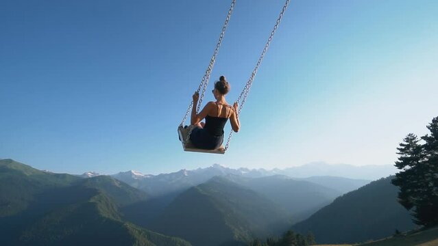 Young woman rides swing over cliff on slope, in front of her background with mountains of Georgia. Tourist place in Khishkili. Easy to fly over mountains carefree. Slow motion
