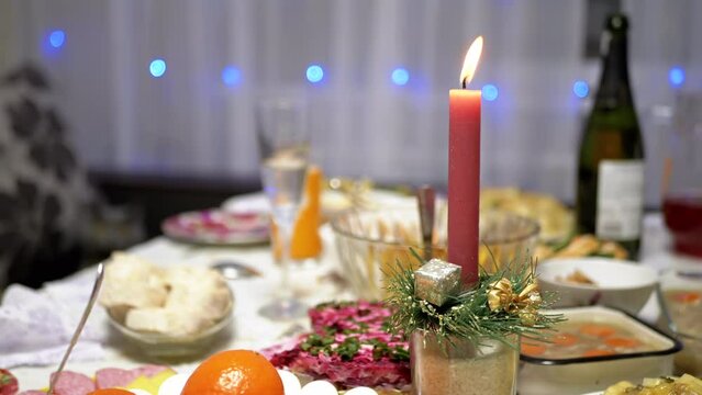 Burning Red Christmas Candle Burns with a Bright Flame on the New Year's Table. Christmas decorated table on eve of holiday on background bulbs garlands. Cozy family dinner in a festive atmosphere.