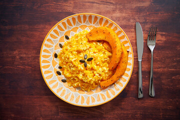 Pumpkin risotto with pumpkin slices photographed from above on a wooden table. Pumpkin seeds as decoration. Knife and fork next to the plate. Italian recipe.