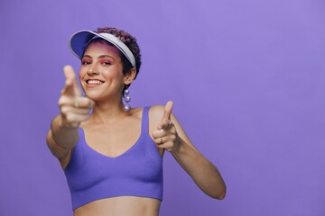 Portrait of a sporty fashion woman posing smiling with teeth and pointing a finger at the camera in a purple yoga tracksuit and a transparent cap on a purple monochrome background