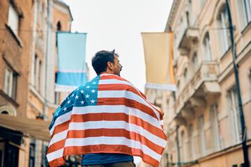 Rear view of sports fan covered with American national flag walk on street.