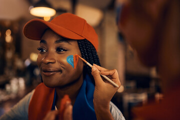 Black soccer fan getting her face painted in colors of her favorite team during world championship.