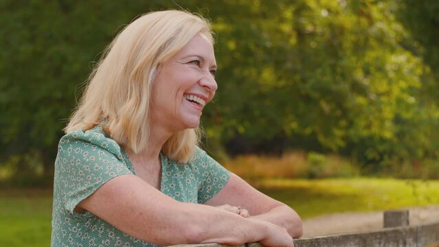 Portrait of laughing mature or senior woman leaning on wooden fence on walk in summer countryside - shot in slow motion