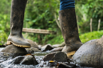 close-up of a hiker girl crossing a stream in black marsh boots. peasant girl stepping on the stones to avoid falling into the water. green background of rain forest in summer.