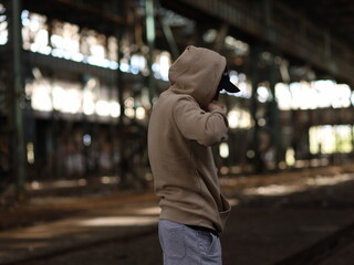a young rapper guy in a hood and cap black jacket in an abandoned old building - obrazy, fototapety, plakaty