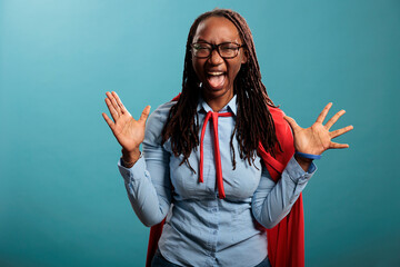 Scared african american superhero woman screaming mouth wide open at camera while standing on blue...