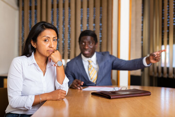 Frustrated hispanic woman entrepreneur sitting at office desk on background with angry partner...