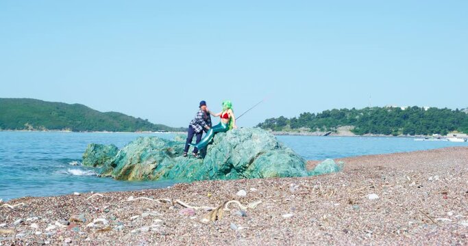 Lucky fisherman in a panama hat and plaid shirt caught a mermaid, sat her down on a rock next to him and wanted to kiss her, but she slapped him in the face, so man fell into the sea