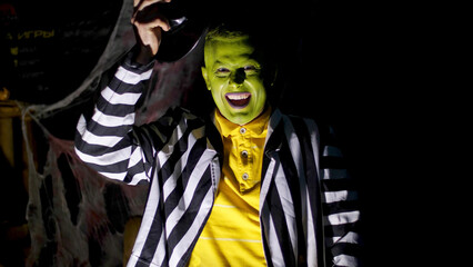 Fototapeta na wymiar Halloween party, night, twilight, in the rays of light, a man with a terrible make-up, with a green face lifts his hat, laughs eerily and runs away. Behind the background is a spiderweb. High quality