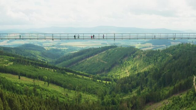 Suspension Sky Bridge with tourists over canyon. Attraction connects hills on spacious highland with forests and curved roads in Dolni Morava aerial view
