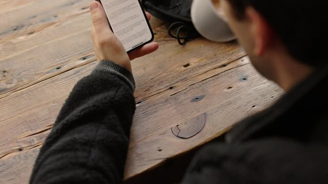 Unrecognizable male takes a sip warm cup of coffee reading news article and surfing internet resources while sitting at the wooden desk in coffee shop in the center of the city 4k