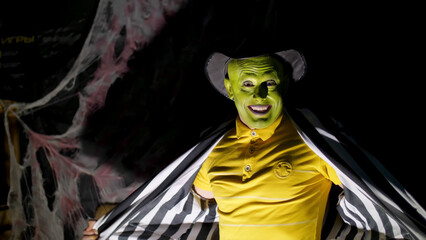 Halloween party, night, twilight, in the rays of light, a man with a terrible make-up, with a green face and a hat laughs eerily, frightens, Behind the background is a spiderweb. High quality photo