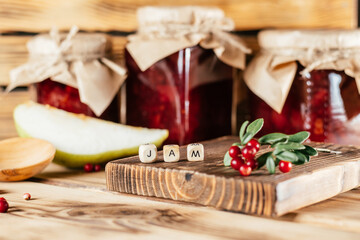 Jars of homemade lingonberry and pear jam with craft paper on lids on wooden surface next to fresh lingonberries and pears.