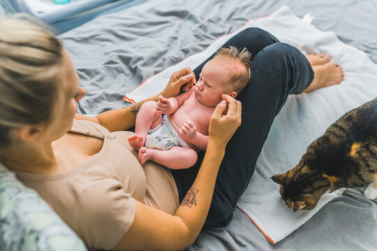 Young mother sitting on a bed, holding her newborn son with cute brown hair on her lap, communicating with him. Big multicolored cat sitting near them and sniffing the area. High quality photo
