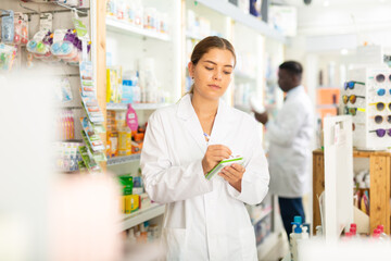 Fototapeta na wymiar Female pharmacist standing in salesroom of drugstore and holding notebook in hands