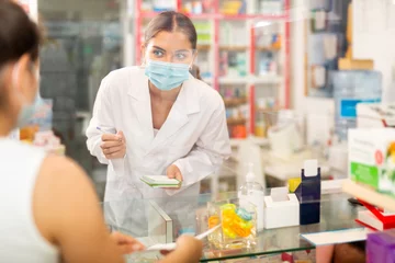 Zelfklevend Fotobehang Young confident female pharmacist in a protective mask, working in a pharmacy during the pandemic, takes an order for a medicine from a buyer © JackF