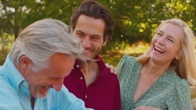 Smiling mature parents with mixed race adult son leaning on wooden fence on walk in summer countryside talking together - shot in slow motion