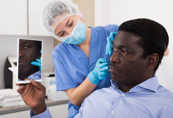 African american male client looking at mirror while qualified beautician examining his face after...