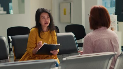 Asian women talking about medical report papers in facility lobby, filling in checkup form before attending exam appointment with physician. Sitting in waiting room at hospital clinic.