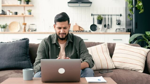 Frustrated sad arabian or indian young man in stylish casual clothes, freelancer, web designer, sit in the living room on the sofa, working on a laptop on a project, upset with results of work or news