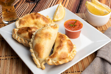 Argentinian homemade empanadas on square white plate red sauce and lemon slices on wooden table