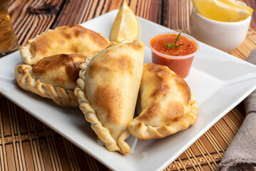 Argentinian homemade empanadas on square white plate red sauce and lemon slices on wooden table