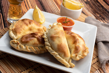 Argentinian homemade empanadas on square white plate red sauce and lemon slices on wooden table