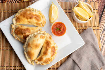 Argentinian homemade empanadas on square white plate red sauce and lemon slices on wooden table