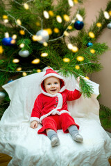 little child baby boy sitting in chair near christmas tree in living room in red santa claus costume