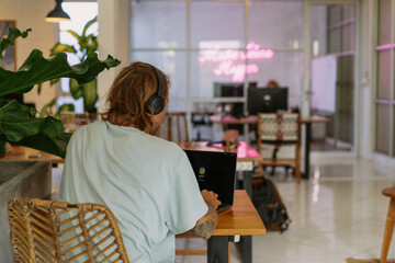 Fototapeta na wymiar A young smiling man in big headphones works on a laptop in a Bali coworking space. Daily call with the team. Work on your laptop from anywhere.