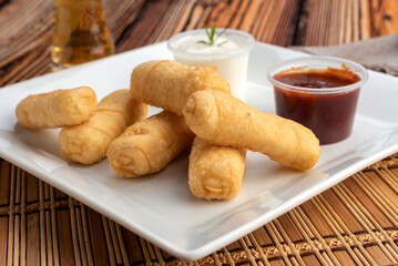 Tequeño sandwiches of dough and cheese with white sauce and red sauce on square white plate on wooden table