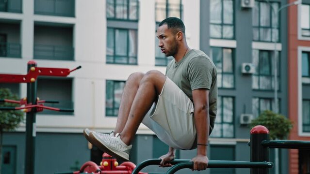 Muscular latino man training abdominal press by lifting legs with bended knees on parallel bars pumping abs muscles at outdoor gym athletic guy sportsman performing masculine bodybuilding exercises